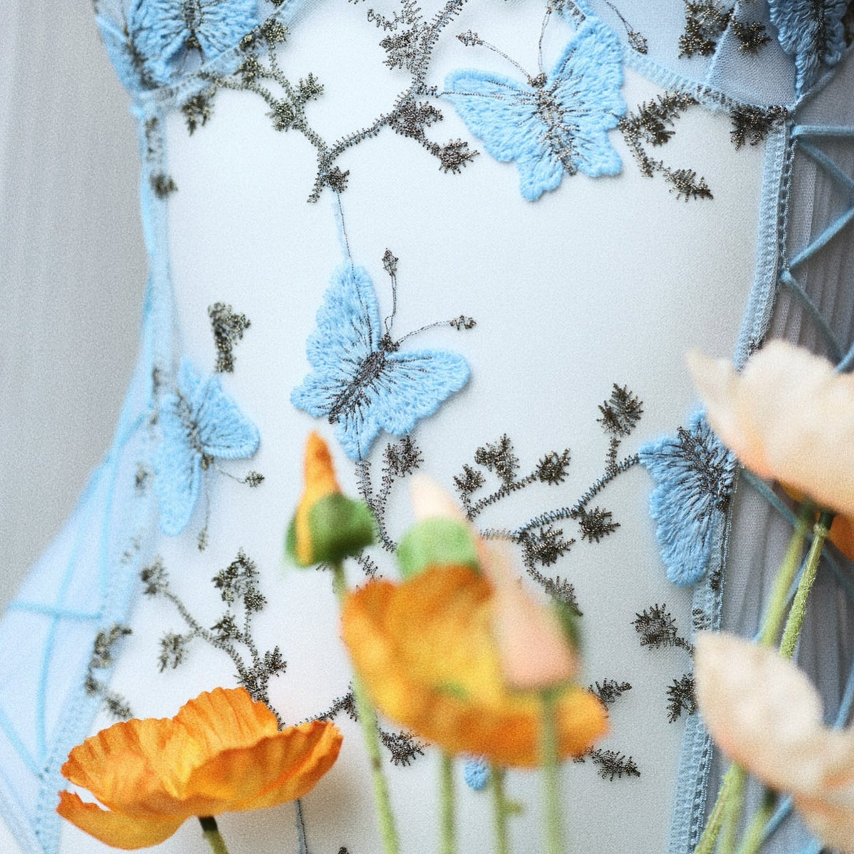 Close-up of delicate blue butterfly embroidery on a sheer bodysuit, showcasing intricate floral details and fine stitching on transparent mesh fabric. Add a touch of elegance to your lingerie collection with Hueslife’s Butterfly Embroidery Bodysuit. The Q00801 features intricate butterfly embroidery, perfect for both comfort and allure.